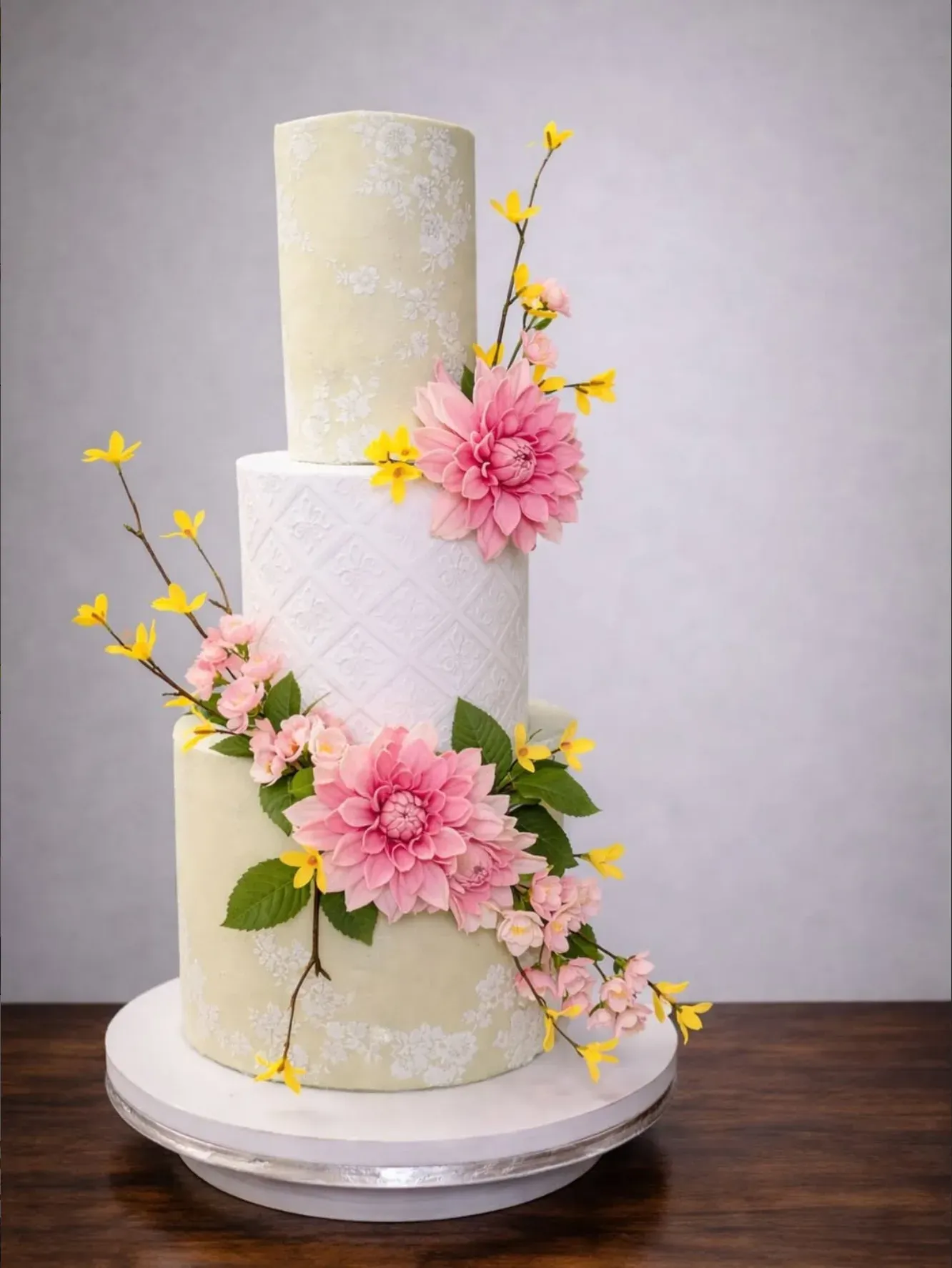 Three-tier wedding cake in sage green and white with lace-effect piping, trimmed with pink sugar dahlias, cherry blossom and sprays of yellow forsythia.